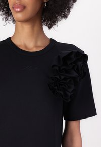 Camiseta de algodón negra con un acento de tela con volantes en el hombro izquierdo y un sutil logo bordado en el pecho. Mangas cortas.