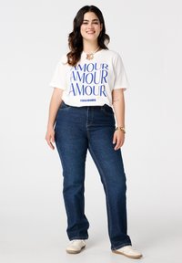 Witte t-shirt met blauwe tekst "AMOUR" en "TOUJOURS." Gecombineerd met donkerblauwe jeans en witte sneakers. Eenvoudige, casual outfit.