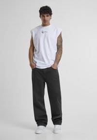 Hvit ermeløs bomullstopp med svart brodert logo, svarte løstsittende jeans og hvite joggesko. Modellen står med hendene i lommene.