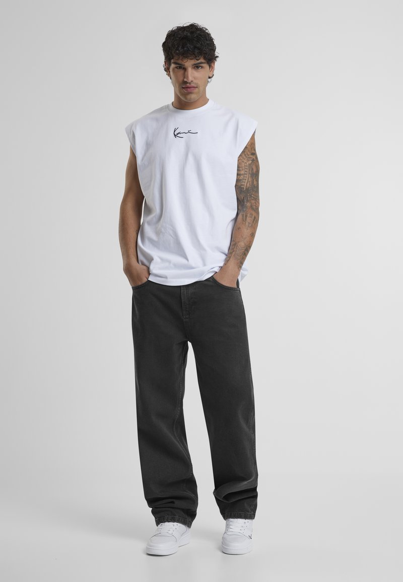 Hvit ermeløs bomullstopp med svart brodert logo, svarte løstsittende jeans og hvite joggesko. Modellen står med hendene i lommene.
