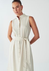 Bezrukávové béžové šaty s vyšším golierom, zapínaním na gombíky a opaskom zviazaným v páse, vyrobené z ľahkého materiálu.