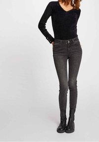 Schwarzer langärmliger Pullover mit V-Ausschnitt und Knopfakzenten, kombiniert mit dunkelgrauen Skinny-Jeans und schwarzen Stiefeletten. Einfaches, figurbetontes Design.