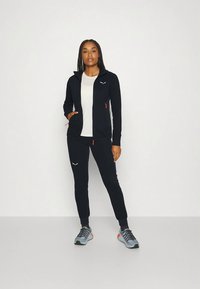 Sieviete stāv melnā kapučjaka, melnās sporta biksēs, baltā krekla un pelēkās kedās pret vienkāršu gaišu fona.