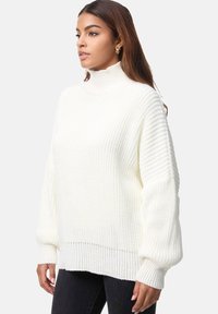 Wijd oversized wit gebreide trui met een hoge kraag, geribbelde structuur, verlaagde schouders en brede mouwen, gecombineerd met zwarte spijkerbroek.