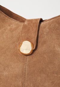 Un matériau en daim brun avec un accent en métal doré lisse et des détails de couture visibles. Le design met en valeur une forme contemporaine et minimaliste.