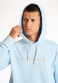 Sudadera azul claro con un bolsillo frontal y capucha con cordón. Presenta "COPENHAGUE LINDBERGH" en texto beige en el pecho. Textura suave.