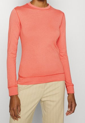 Femme portant une chemise corail ajustée à manches longues et un pantalon beige à fines rayures, debout devant un fond uni.