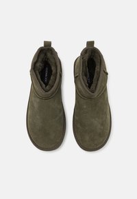Paire de chaussons en daim vert olive à enfiler, doublés d'une fourrure sombre moelleuse, avec des languettes au talon, vus de dessus sur fond blanc.