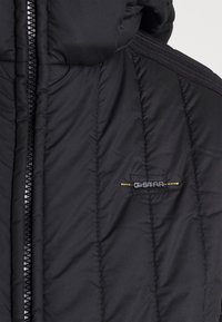 Gros plan sur une veste matelassée noire avec une fermeture éclair frontale et un petit logo G-Star brodé sur la poitrine.