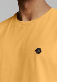 Geel katoen t-shirt met een ronde halslijn en een zwart hexagonaal logo-detail op de borst. Zachte textuur, eenvoudig ontwerp.