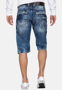 Shorts en denim bleu avec une texture délavée, coupe mi-genou, deux poches arrière et une marque sur la poche droite. Portés avec des baskets blanches.