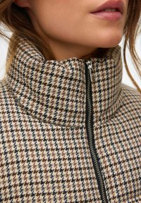 Sako s kostkovaným vzorem houndstooth v béžové, hnědé a černé barvě, s vysokým límcem a zipovým zapínáním u výstřihu.