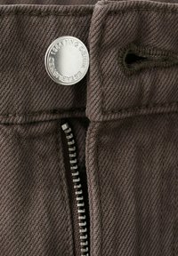 Primer plano de tela de mezclilla marrón con un botón metálico que dice "MNG DENIM ESTABLISHED 1984" y una cremallera parcialmente visible.