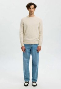 Camisola de malha bege claro com decote redondo e punhos canelados, combinada com jeans azul claro de perna reta e sapatos pretos com detalhes brancos.