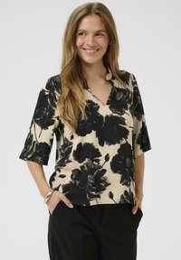 Moteris šypsosi, vilkinti smėlio spalvos bluzoną su dideliu juodu gėlėtu raštu ir juodomis kelnėmis, rankos kišenėse, prieš vientisą foną.