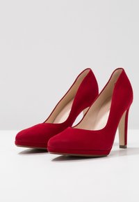 Røde ruskinds højhælede sko med spids tå og en glat, mat overflade. Har en slank stiletto-hæl og en platform base.