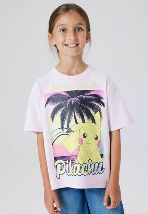 Ragazza sorridente che indossa una maglietta rosa chiaro con grafica di Pikachu e palme, abbinata a jeans blu, in piedi davanti a uno sfondo semplice.