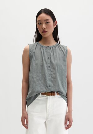 Jeune femme portant un chemisier gris sans manches boutonné, rentré dans un pantalon blanc avec une ceinture marron, debout devant un fond blanc uni.