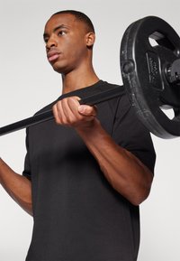 Mann in schwarzem Hemd hält eine Langhantel mit Gewichtsscheiben auf Schulterhöhe und bereitet sich auf das Heben vor, vor einfachem Hintergrund.