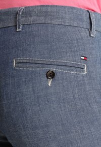 Gros plan sur la poche d'un pantalon bleu avec un bouton et un petit logo de marque rouge, blanc et bleu marine en haut à droite.