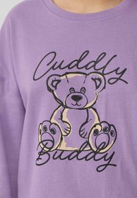 Lavendelkleurige sweatshirt met een zwarte omtrektekening van een beer en de tekst "Cuddly Buddy" in cursief onderaan. Zachte stoftextuur.