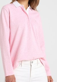 Helles rosa Langarm-Polohemd mit weißem Kragen, featuring einen lässigen Schnitt und subtiler Struktur. Getragen mit weißen Hosen, die Details beider Bekleidungsstücke zeigen.