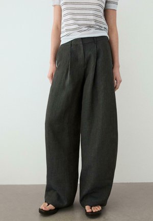 Personne portant un pantalon large gris foncé, un haut à manches courtes rayé blanc et noir, et des sandales noires, debout à l'intérieur.