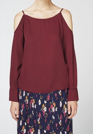 Burgunderfarbene Kalt-Schulter-Bluse mit langen Ärmeln und lockerer Passform, aus einem glatten Stoff gefertigt; kombiniert mit einem floral bedruckten, plissierten Rock in dunklem Marineblau.