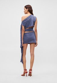 Σετ δύο τεμαχίων από σκουρό μπλε ύφασμα. Τοπάνι με έναν ώμο και ρευστή λεπτομέρεια· μίνι φούστα. Το μοντέλο φορά κόκκινες ψηλοτάκουνες σανδάλες με λουράκια.