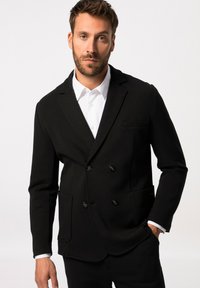 Blazer negro de doble botonadura en tejido elástico, con solapa con muesca, bolsillos frontales y botones negros. Debajo, se ve una camisa blanca.