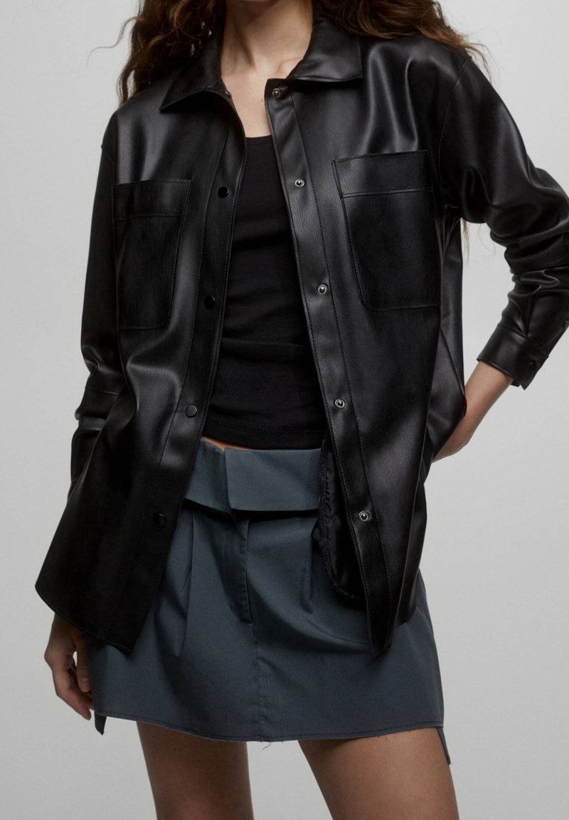 Black faux leather shirt jacket with a collared neckline, front pockets, and snap buttons, paired with a gray pleated skirt.