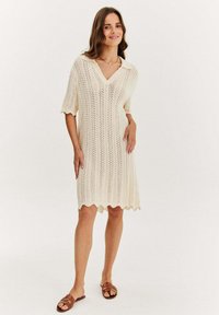 Sukienka dzianinowa w kolorze kremowym z dekoltem w serek i krótkimi rękawami, z teksturowanym wzorem, falbaniastym dołem, w zestawie z brązowymi sandałami na płaskiej podeszwie.