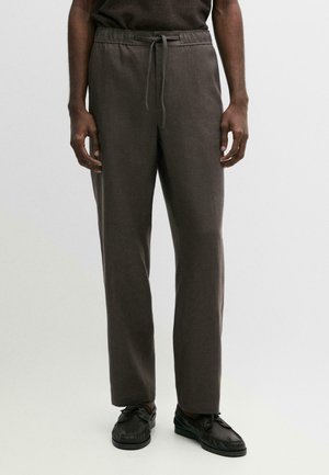 Mann trägt dunkelbraune gerade geschnittene Hose mit elastischem Bund und Kordelzug, kombiniert mit schwarzen Schnürschuhen, steht auf weißem Boden.