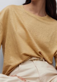 Kvinna i en lös senapsgul topp instoppad i högmidjade krämfärgade byxor med ett beige bälte och guldspänne, sittande mot en neutral bakgrund.