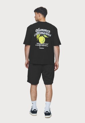 Fekete pamut póló citrom grafikával és szöveggel a hátsó részen, fekete sorttal és kék sportcipővel, fehér kiegészítőkkel.