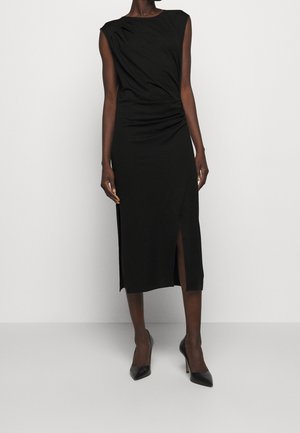 Mujer con un vestido midi negro sin mangas con una abertura lateral y detalle fruncido, combinado con zapatos de tacón alto negros puntiagudos sobre un fondo liso.