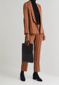 Braunes Nadelstreifenanzug mit schwarzem Rollkragenpullover, hält eine schwarze Lederhandtasche. Hervorgehoben durch spitze Stiefeletten und einen strukturierten Blazer.