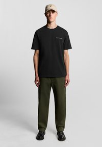 Camiseta de algodón negra de manga corta, combinada con pantalones verde oliva. Se lleva una gorra beige con un logo, complementada por zapatos negros con cordones.