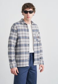 Καρό πουκάμισο σε αποχρώσεις του γκρι και μπλε, με κουμπιά μπροστά, τσέπη στο στήθος και μακριά μανίκια. Φορεμέ νο πάνω από λευκό μπλουζάκι και μπλε σκούρο παντελόνι.