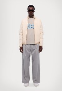 Lichtcreme pufferjack met een grijze graphic t-shirt met blauwe print, brede lichtgrijze broeken, witte sneakers en zonnebril.