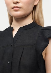 Femme à la peau claire et aux cheveux blonds portant un chemisier noir sans manches avec des boutons et un col arrondi.