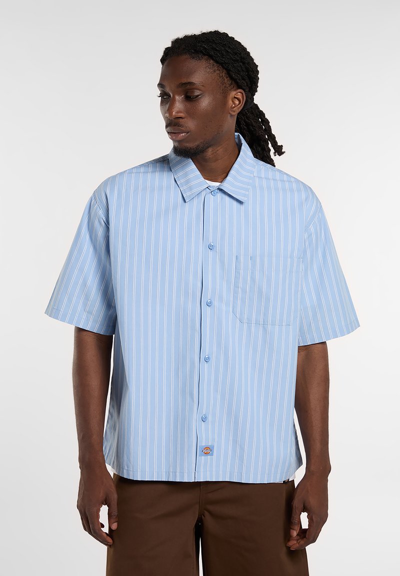Kortærmet skjorte med knapper i lyseblå med hvide lodrette striber, der har en brystlomme og et diskret logomærke nær kanten.