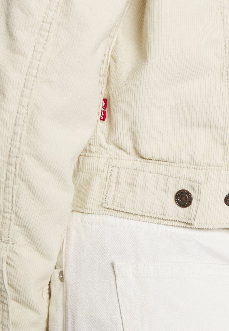 Veste en velours côtelé beige avec une étiquette rouge de Levi's, des boutons-pression marron, et une finition texturée. Accents en denim blanc en bas.