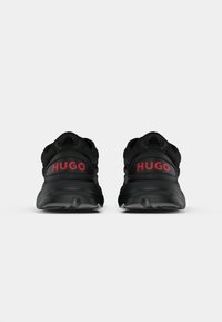 Sorte treningssko med strukturert gummisåle og polstret krage, med "HUGO" i rødt på hælen og en strømlinjeformet design.