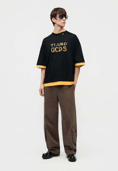 Junger Mann trägt ein übergroßes schwarzes T-Shirt mit gelbem Besatz und goldenem Schriftzug, braune Cargo-Hose, schwarze Schuhe und große dunkle Sonnenbrille.