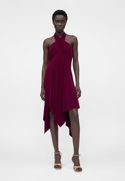 Γυναίκα με αμάνικο ασύμμετρο μπορντό φόρεμα με λαιμόκοψη halter, σε συνδυασμό με μπεζ πέδιλα με ψηλό τακούνι, στέκεται μπροστά σε απλό φόντο.