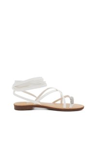 Sandali bianchi con cinturini, dalla suola piatta, con un fermaglio adornato da cristalli e cinture sottili. Il materiale sembra liscio e flessibile.