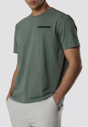 Uomo con maglietta verde oliva a maniche corte con piccolo logo nero sul petto, mani nelle tasche di pantaloni chiari, in piedi davanti a uno sfondo semplice.