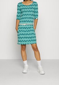 Turkusowa sukienka z wzorem w zygzaka, z rękawami do trzech kwart, posiadająca kieszenie; z białym paskiem w talii, noszona w białych trampkach.