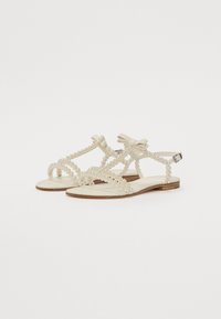 Beige tasapohjaiset sandaalit, joissa on helmiä muistuttavia koristeita, koristeellisia nauhoja ja hihnadesign. Pohja on valmistettu vaaleanruskeasta materiaalista.